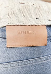 Beige strikket stof med et tekstureret mønster øverst, der har et lysebrunt lædermærke præget med "RETERNITY," syet på denim.