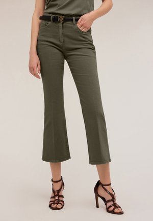 Vrouw draagt olijfgroene cropped broek met zwarte riem en donkerbruine sandalen met hoge hakken, staand met één hand in haar zak.