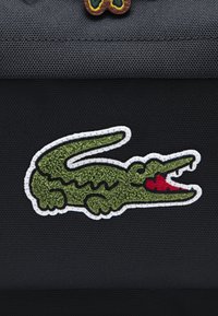 Grünes, besticktes Krokodil-Logo mit roten Details auf einem strukturierten schwarzen Stoffhintergrund, umrandet von weißer Naht.