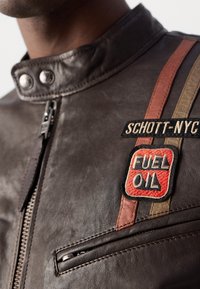 Gros plan d'une veste en cuir marron avec fermeture éclair à l'avant, col boutonné, poche zippée et écussons ornés des inscriptions "SCHOTT-NYC" et "FUEL OIL."