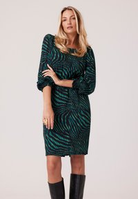 Sukienka z zielono-czarnym wzorem w paski zebry, długimi bufkami, okrągłym dekoltem oraz wysmukloną sylwetką, zestawiona z czarnymi kozakami za kolano.