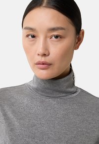 Kobieta o prostych, ciemnych włosach zaczesanych do tyłu, ubrana w szarą, błyszczącą bluzkę z wysokim kołnierzem, patrzy prosto w kamerę z neutralnym wyrazem twarzy.