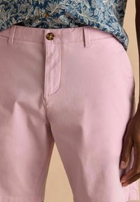 Helle rosa Shorts aus weichem Stoff, mit Knopfverschluss und schrägen Taschen, kombiniert mit einem blauen Hemd mit Blumenmuster.