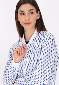 Jachetă bomber albă cu model abstract albastru, închidere cu fermoar frontal, guler și mansete albe contrastante, țesătură ușoară, croi relaxat.