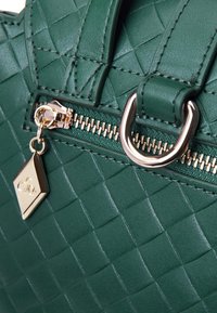 Borsa a tracolla verde texturizzata con motivo trapuntato, caratterizzata da una cerniera dorata, un dettaglio in metallo circolare e un piccolo charm a forma di etichetta dorata.