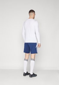 Langärmliges weißes Sportshirt, blaue Shorts mit weißen Seitenstreifen, knielange weiße Socken mit schwarzen Streifen und schwarze Fußballschuhe.