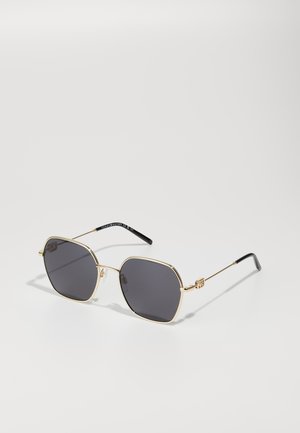 Gold-framed octagonal sunglasses with dark gray lenses. Features thin metal arms and a black temple tip. Sleek, modern design.