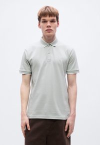 Ανοιχτό γκρι πόλο πουκάμισο από βαμβάκι, με κλασικό γιακά, δύο κουμπιά και μικρό λογότυπο στον αριστερό θώρακα. Λεπτή υφασμάτινη υφή.