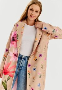 Béžový kabát s kvetinovým vzorom vyrobený z hladkej tkaniny, zdobený ružovými, fialovými a oranžovými kvetmi, skombinovaný s bielym vrchom a džínsovými nohavicami.