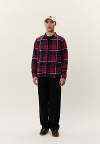 Ung mand iført beige baseballkasket, rød og navy ternet skjorte med knapper, sorte bukser og brune sko, stående mod en ensfarvet baggrund.