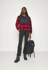 Červeno-čierny károvaný fleeckový pulóver s prešívanou čiernou vestou, čierne nohavice a čierny batoh s emblémom, obutý s čiernymi topánkami.