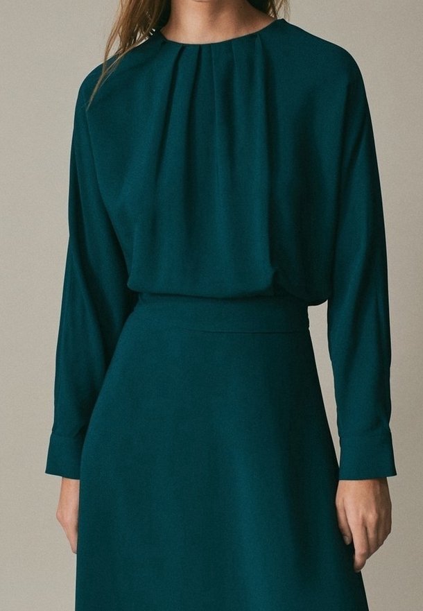 Donna che indossa un abito lungo teal con pinces sullo scollo e una vita aderente su uno sfondo semplice.