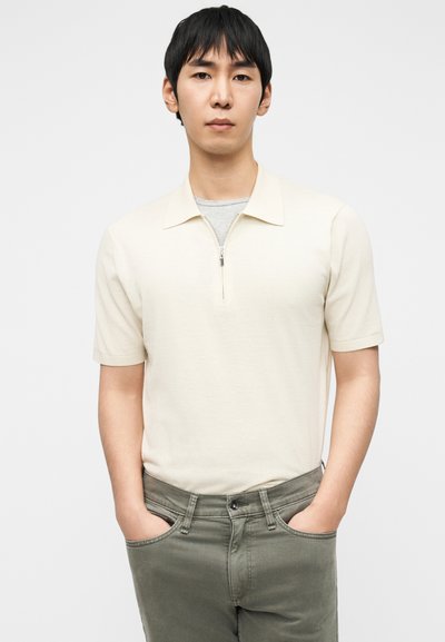 Ανοιχτό μπεζ κοντομάνικο πόλο με φερμουάρ στον γιακά. Φορεμένο πάνω από γκρι κοντομάνικο μπλουζάκι. Συνδυάζεται με παντελόνι τζιν σε ελιά πράσινη απόχρωση.