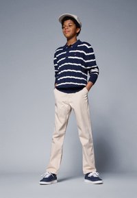 Marineblå og hvite stripete strikkegenser, beige bukser og marineblå sneakers. Grå caps. Modellen står med hendene i lommene mot en nøytral bakgrunn.