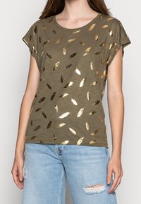 Camiseta verde olivo de manga corta, con patrones de hojas doradas metálicas y cuello redondo, combinada con jeans claros desgastados.
