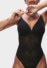 Body in pizzo nero con scollo a V profondo, rifinitura decorativa smerlata, motivo floreale e apertura alta sulle gambe. Tessuto liscio ed elastico.
