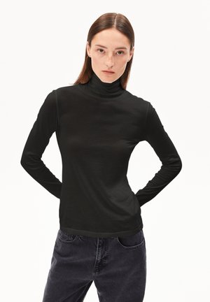 Zwarte longsleeve coltrui van een gladde, rekbare stof. Kenmerkend zijn de getailleerde mouwen en de hoge kraag, gecombineerd met donkerdenim jeans.