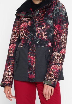 Personne portant une veste multicolore à motifs avec des tons noirs et rouges et des poches noires unies, associée à un pantalon rouge.