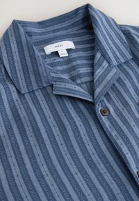 Blauw overhemd met verticale streepjespatroon in verschillende texturen, korte kraag en een enkele bruine knoop als detail.