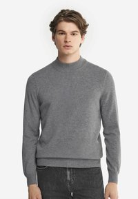 Grauer Strickpullover mit hohem Kragen, langen Ärmeln und gerippten Bündchen. Glatte Textur, taillierter Schnitt, kombiniert mit dunklen Jeans.