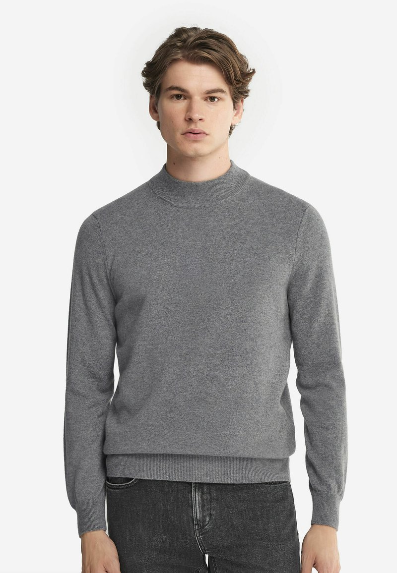 Grauer Strickpullover mit hohem Kragen, langen Ärmeln und gerippten Bündchen. Glatte Textur, taillierter Schnitt, kombiniert mit dunklen Jeans.