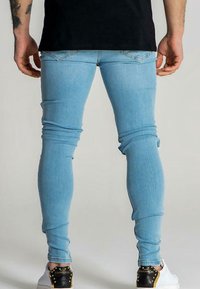 Hombre con jeans ajustados de color azul claro y zapatillas blancas con detalles en negro y dorado en el tacón, visto desde atrás.