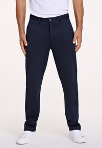 Pantaloni su misura blu navy realizzati in tessuto liscio, con chiusura a bottone e tasche laterali, abbinati a scarpe da ginnastica bianche.