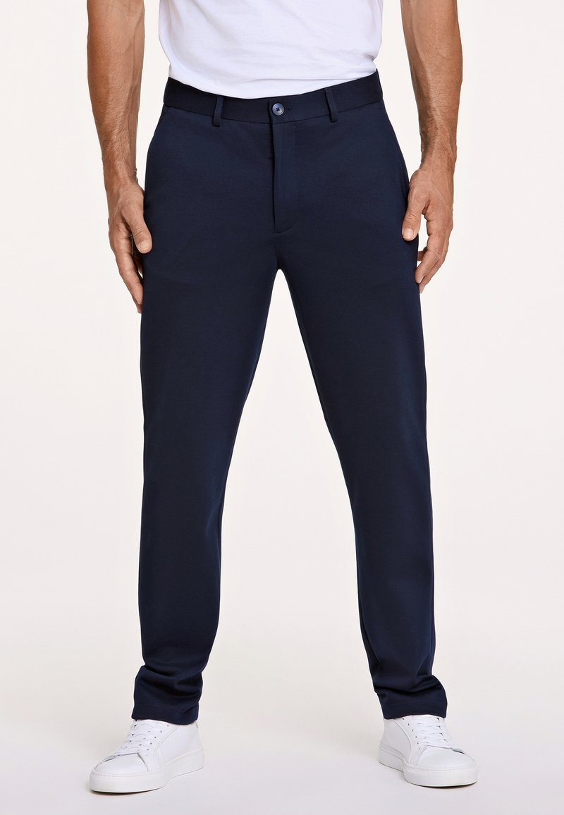 Pantaloni su misura blu navy realizzati in tessuto liscio, con chiusura a bottone e tasche laterali, abbinati a scarpe da ginnastica bianche.