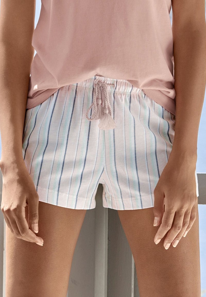 Femme portant un haut sans manches rose clair et un short blanc avec des rayures verticales bleues et roses, debout, les bras détendus le long du corps.