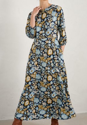 Kobieta ubrana w długą sukienkę maxi z długim rękawem w niebieskie, białe i musztardowe kwiatowe wzory, w parze z brązowymi botkami za kostkę.