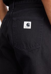 Black jeans featuring a back pocket with a white square label displaying a stylized logo. The fabric has a smooth, sturdy texture.
