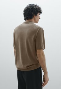 Polo shirt a maniche corte, marrone, in un morbido tessuto a maglia, con colletto classico e vestibilità rilassata, abbinato a pantaloni scuri.