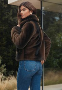 Pruun faux karvane jakk kõrge kraega, mille juurde on kombineeritud sinised teksapüksid. Jakk on pehme tekstuuriga ja lühikese disainiga.