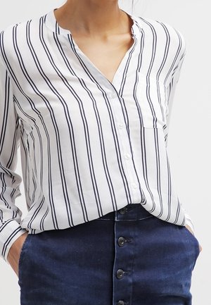 Biała koszula na guziki z pionowymi granatowymi paskami, kołnierzem typu mandarynka oraz kieszonką na piersi, zestawiona z granatową spódnicą z guzikami.