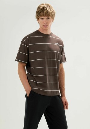 Junger Mann mit kurzem blondem Haar steht, trägt ein braun gestreiftes T-Shirt und schwarze Hose, Hände in den Taschen, neutraler Hintergrund.