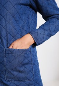 Quiltad blå denimjacka med diamantmönster, långa ärmar och en framficka på vänster sida med synliga sömnadsdetaljer.