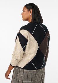 Sweter dzianinowy w beżowym, czarnym i brązowym wzorze argyle z białymi akcentami; ma rękawy o długości 3/4 i ściągacz na dole.