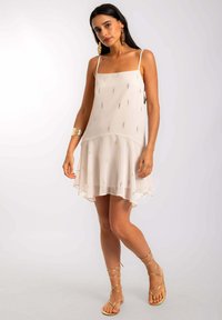Vestido blanco sin mangas con escote cuadrado, dobladillo en capas y acentos de hilo metálico. Combinado con sandalias doradas con tiras y joyería.