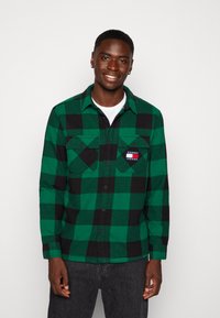 Camicia in flanella a quadri verde e nera con maniche lunghe, bottoni neri, due taschini sul petto e una toppa con logo sulla tasca sinistra.
