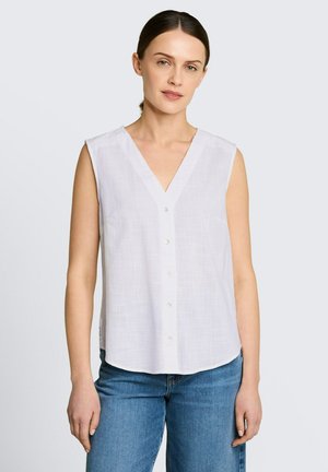 Femme aux cheveux foncés attachés, portant une blouse blanche sans manches boutonnée et un jean bleu, debout devant un fond uni.