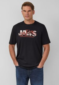 Schwarzes Baumwoll-T-Shirt mit einem Grafikdruck des Wortes "JAWS" in Rot, ergänzt durch detaillierte Bilder und darunter stehenden Text. Rundhalsausschnitt, kurze Ärmel.