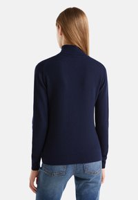 Laivastonsininen neulepusero, valmistettu pehmeästä neuloksesta, jossa on korkea kaulus, pitkät hihat ja vartalonmyötäinen malli, jossa resorit hihansuissa ja helmassa.