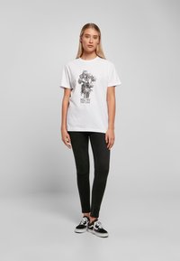 Hvit bomull t-skjorte med svart blomsterdesign og teksten "VENT IKKE TIL DET ER FOR SENT." Sammen med svarte denimjeans og svarte joggesko.