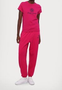 Henkilö, joka on pukeutunut kirkkaan pinkkiin Ralph Lauren -t-paitaan ja vastaaviin verkkareihin, valkoiset lenkkarit jalassa, seisoo yksinkertaista valkoista taustaa vasten.