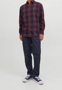 Camicia a quadri bordeaux e blu navy con maniche lunghe, abbinata a pantaloni blu navy e sneakers blu navy con lacci bianchi.