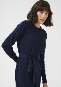 Robe en tricot bleu marine à manches longues, col rond et taille ceinturée. Texture douce avec un accent de ceinture nouée à la taille.