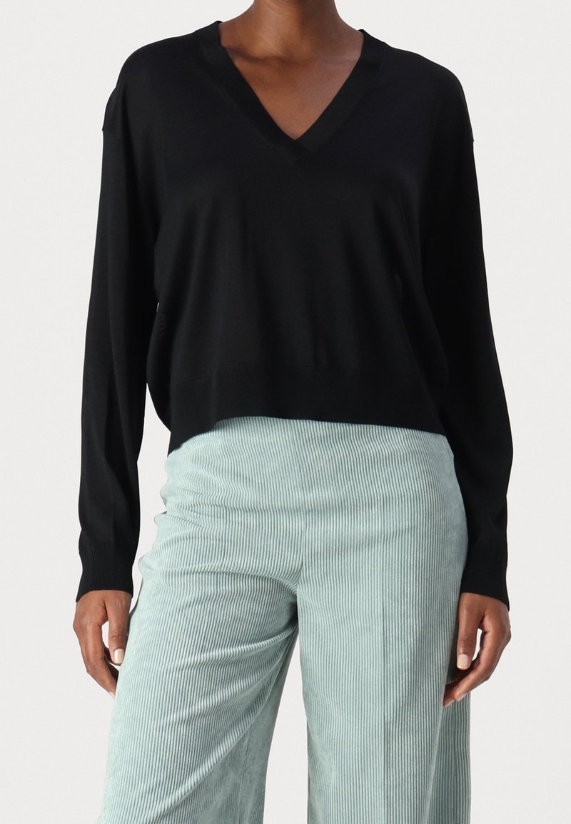 Haut noir à manches longues avec col en V, associé à un pantalon large côtelé taille haute bleu clair, porté par une personne debout.