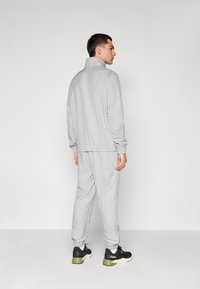 Lys grå sportsdragt med elastiske manchetter og talje, parret med sorte og grønne sneakers, set fra ryggen.