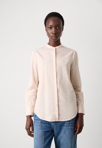 Blusa rosa chiaro a maniche lunghe con colletto a banda, chiusura frontale con bottoni e orlo curvo. Tessuto morbido con una vestibilità leggermente strutturata.