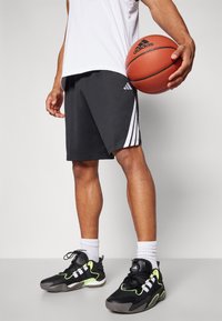Čierne atletické šortky s bielymi pruhmi a logom Adidas, v kombinácii s čiernymi a neonovo zelenými topánkami. Hráč drží basketbal.